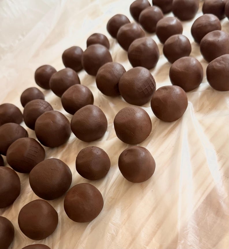 K&ouml;nnte beinhalten: Nahaufnahme von zahlreichen dunkelbraunen, kugelf&ouml;rmigen Objekten, die Tr&uuml;ffeln oder Pralinen &auml;hneln, auf einer wei&szlig;en Oberfl&auml;che. Einige Kugeln weisen strukturierte Muster auf. Die Beleuchtung betont die glatte Textur der Leckereien.