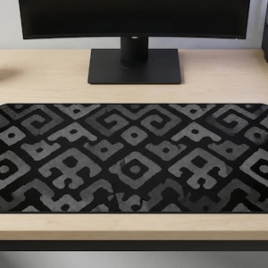 Puede incluir: Una alfombrilla de escritorio con estampado negro y gris con un diseño geométrico. La alfombrilla es rectangular y se encuentra sobre un escritorio de madera de color claro. Un monitor de ordenador, una pequeña planta en maceta y un portalápices también están en el escritorio.