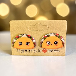May include: A pair of handmade taco-shaped earrings on a brown card. The earrings feature a smiling taco design with orange, red, and green details. The card reads "Handmade with love."