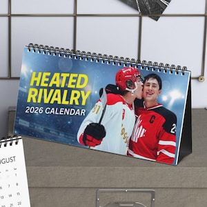 May include: Two desk calendars. One calendar shows a photo of two shirtless men and the month of August. The other calendar has the text "HEATED RIVALRY 2026 CALENDAR" and a photo of two hockey players. The calendars are on a desk.