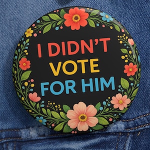 May include: Round button with the text "I DIDN'T VOTE FOR HIM" in red, yellow, and blue, surrounded by a floral wreath of red, pink, yellow, and blue flowers and green leaves. The button is set against a denim jacket.