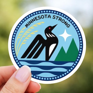 Puede incluir: Adhesivo redondo con un diseño que presenta un somorgujo negro y blanco, montañas verdes y una estrella blanca sobre un fondo azul claro. El texto "MINNESOTA STRONG" está dispuesto en círculo.