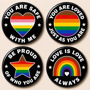 Puede incluir: Cuatro botones redondos negros con diseños de arcoíris y texto blanco. Los botones tienen frases como "Estás a salvo conmigo" y "El amor es amor siempre". Cada botón tiene un diseño diferente, incluyendo un corazón, un círculo, una estrella y un arcoíris.