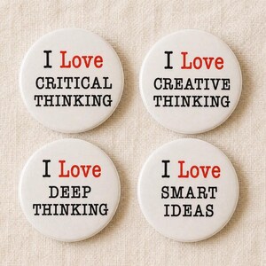 May include: Four white circular badges with the words "I Love" in red, followed by "CRITICAL THINKING", "CREATIVE THINKING", "DEEP THINKING", and "SMART IDEAS" in black. They are displayed on a beige fabric.