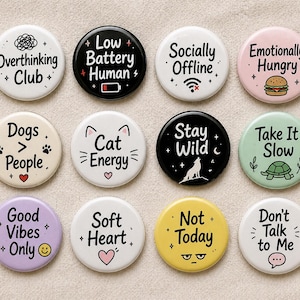 Op de afbeelding: Een verzameling van twaalf ronde buttons met verschillende zinnen en afbeeldingen. De buttons bevatten teksten als "Overthinking Club", "Low Battery Human" en "Dogs > People". Kleuren zijn wit, zwart, roze, groen, geel en paars.