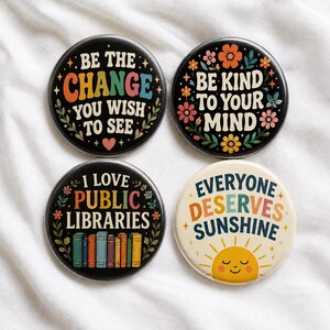 May include: Four circular pinback buttons with text and graphics. Three buttons have black backgrounds with floral accents. One button has a cream background with a yellow sun. Text includes: Be the change you wish to see, Be kind to your mind, I love public libraries, and Everyone deserves sunshine.