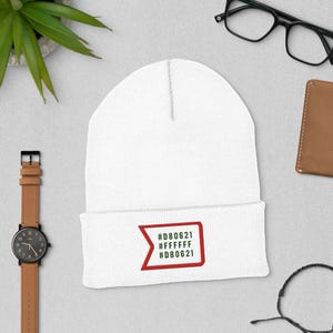 May include: A white beanie with a red and black graphic. The graphic contains the text "#D80621 #FFFFFF #D80621" inside a red rectangle. The beanie is made of a soft material and is suitable for cold weather.