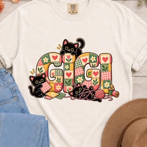 Può includere: Una t-shirt color avorio con una grafica della parola GIGI in un motivo patchwork con disegni di fiori e cuori. Tre gatti neri sono integrati nel design, uno dei quali gioca con un gomitolo di lana rosa e accessori per il cucito.