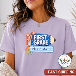 Puede incluir: Una camiseta de color morado claro con un gráfico de un lápiz azul con un lazo a cuadros rojo y blanco, una regla y el texto "FIRST GRADE" y "Mrs. Anderson". Una insignia circular muestra una flor de algodón y el texto "100% COTTON".