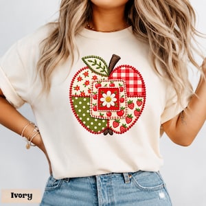 Op de afbeelding: Een ivoorkleurig t-shirt met een grote, centrale appelvormige afbeelding bestaande uit diverse patchwork-stijl patronen, waaronder rood gingham, groene stippen, bloemenprints en kleine aardbeien, met een witte margriet in het midden.
