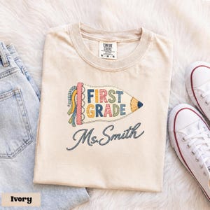 Maglietta personalizzata Comfort Colors® per insegnanti di prima elementare, modello patchwork con matita e bandierina