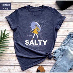Op de afbeelding: Heather marineblauwe t-shirt met een afbeelding van een persoon onder een paraplu, met het woord "SALTY" in witte letters. Het shirt wordt gecombineerd met een blauwe jeans en een zwarte pet. De tekst "All the flavors and you choose to be..." staat er ook op.