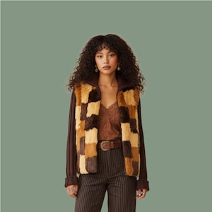 May include: A patchwork faux fur vest in shades of brown and tan, featuring a brown knit trim and a zip. The vest is worn over a brown camisole, brown pinstripe trousers and a brown belt. The model has dark, curly hair.