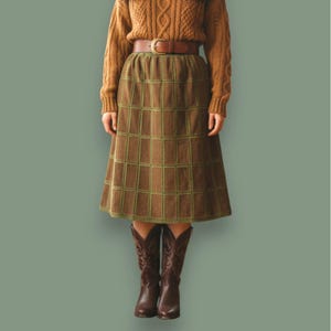 Peut inclure: Une personne porte un pull en tricot torsadé marron, une ceinture marron et une jupe marron avec un motif quadrillé vert. Elle porte également des bottes de cowboy marron. L'ensemble est posé sur un fond vert.