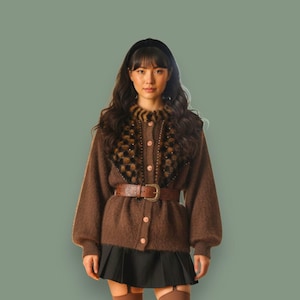 May include: A brown cardigan with a decorative collar, a brown belt, and a black skirt. The cardigan has a textured appearance and is buttoned up. The collar features a patterned design. The outfit is completed with a black headband.