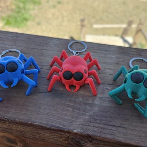 May include: Three colorful spider keychains in blue, red, and teal are displayed on a wooden surface. Each keychain features a spider design with large, round eyes and a metal keyring. The keychains are a fun accessory.
