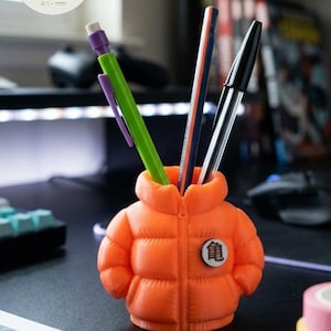 Puede incluir: Un portalápices naranja con forma de chaqueta acolchada, con cremallera y un emblema circular. Contiene bolígrafos y lápices. El logotipo de Atelier La Sylle es visible en el fondo.