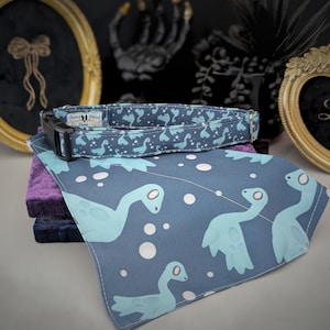 May include: A blue dog collar and matching bandana set. The collar and bandana feature a repeating pattern of light blue Loch Ness Monster illustrations on a dark blue background. The collar has a black buckle and a small tag with text.