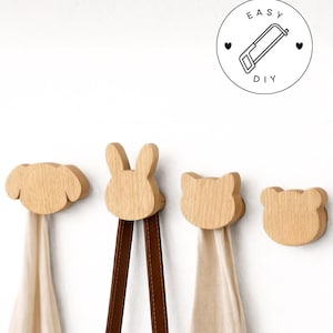 Może przedstawiać: Drewniane haczyki ścienne w kształcie zwierząt: psa, królika, kota i niedźwiedzia. Każdy haczyk jest wykonany z jasnego drewna i trzyma szalik lub torbę. Obraz zawiera logo DIY.