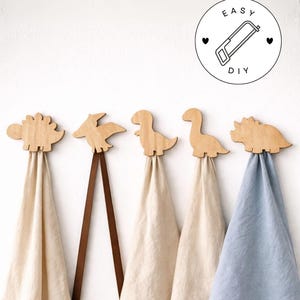 May include: Wooden dinosaur-shaped towel hooks with hanging towels. The hooks are in the shape of a Stegosaurus, Pterodactyl, Tyrannosaurus Rex, Brontosaurus, and Triceratops. The towels are beige and blue. The image includes a DIY logo.