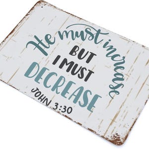 Op de afbeelding: Rechthoekig metalen bord met een verweerde witte achtergrond en bruine randen. Het bord toont de tekst "He must increase but I must decrease" in turquoise en zwarte letters, met "John 3:30" eronder.