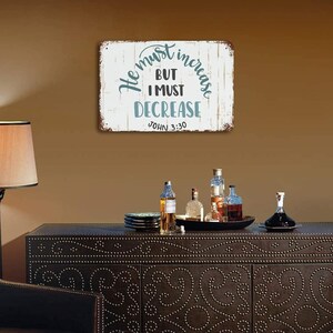 Op de afbeelding: Een rechthoekig metalen bord met de tekst "He must increase, but I must decrease" in turquoise en zwarte letters. Het bord heeft een verweerde, rustieke uitstraling en is op een bruine muur gemonteerd.