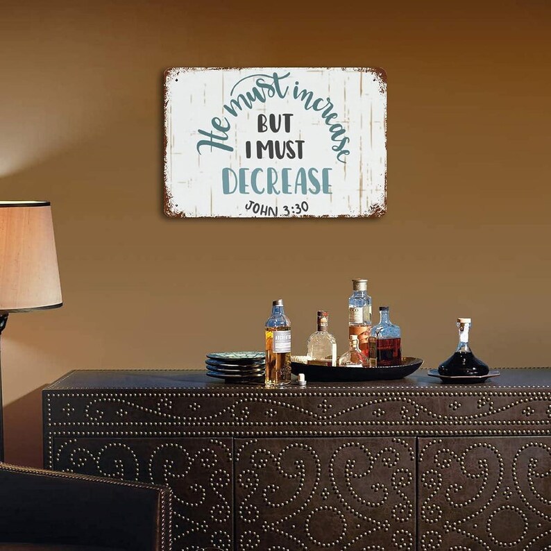 Op de afbeelding: Een rechthoekig metalen bord met de tekst "He must increase, but I must decrease" in turquoise en zwarte letters. Het bord heeft een verweerde, rustieke uitstraling en is op een bruine muur gemonteerd.