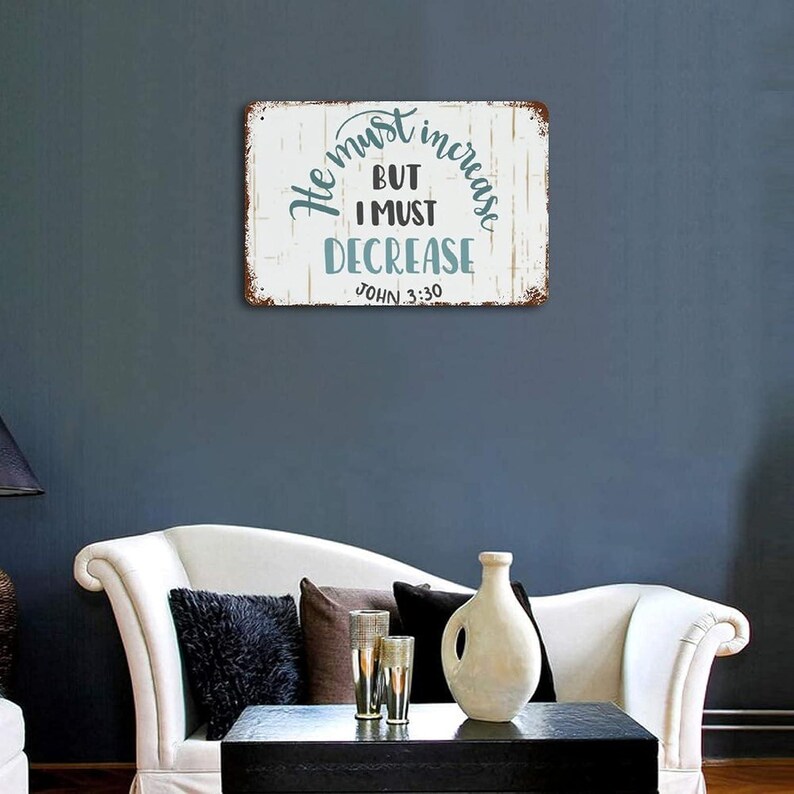 Op de afbeelding: Een rechthoekig metalen bord met een verweerde witte achtergrond en een bruine rand. Het bord bevat de tekst "He must increase, but I must decrease, John 3:30" in turquoise en zwarte letters. Het bord is bevestigd aan een donkerblauwe muur.
