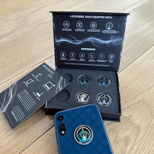 May include: A box set containing six circular stickers with a central design, a blue smartphone with a matching sticker, and a card with suggested uses. The box is black with white text and graphics. The phone has a circular sticker on the back.