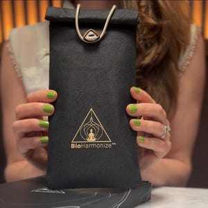 May include: A black pouch with a gold-colored logo and the text "BioHarmonize". The pouch has a cream-colored drawstring and a gold-colored clasp. The pouch is held by a person with green nail polish.