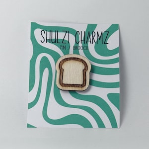 May include: A wooden pin or brooch shaped like a slice of bread. The pin features a light brown centre and a darker brown outline. The pin is displayed on a card with the text "SHULZI CHARMZ PIN / BROOCH" and a teal and white wavy pattern.