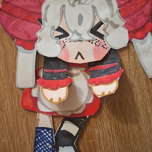 May include: A handmade paper doll with a red and white dress, black and red striped sleeves, and blue shoes. The doll has a white face with freckles and a black triangle on top of its head.