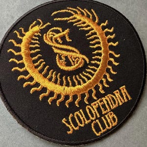 Peut inclure: Écusson rond brodé noir avec un motif doré représentant un scolopendre stylisé et le texte "SCOLOPENDRA CLUB". L'écusson est probablement destiné aux vêtements ou aux accessoires.