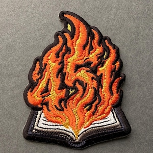 May include: An embroidered patch featuring an open book with white pages and a black cover, topped with stylized flames in vibrant shades of orange, yellow, and red, all outlined with a thick black border on a dark gray background.