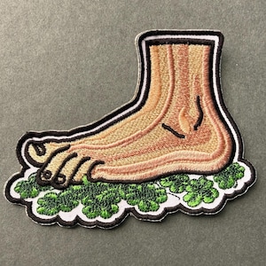 May include: Embroidered patch featuring a foot with a light brown skin tone, resting on a bed of green leaves. The patch has a black and white border. The foot is detailed with lines and shading.