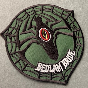 May include: A circular patch with a green background and a black spider design. The spider has a red and yellow eye. The words "BEDLAM BRIDE" are embroidered in white.
