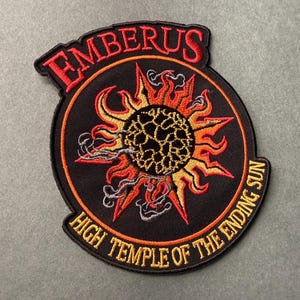 Puede incluir: Parche bordado con la palabra "EMBERUS" en texto rojo arqueado sobre un diseño circular. El diseño presenta un sol estilizado con llamas naranjas y amarillas, y el texto "HIGH TEMPLE OF THE ENDING SUN" en dorado.
