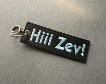 May include: A rectangular black leather keychain featuring the embroidered light blue text Hiii Zev! with a silver-toned metal split ring attached to the left side through a reinforced metal eyelet, showcasing clean stitching along the edges.