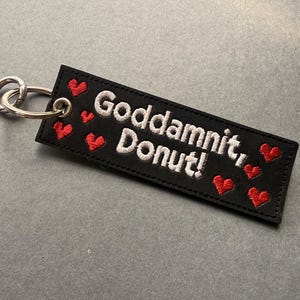 May include: Black rectangular keychain with the embroidered phrase "Goddamnit, Donut!" in white, accompanied by red heart accents. Includes a silver metal keyring.