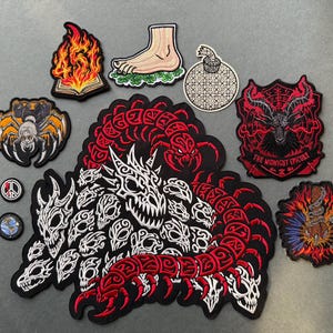 May include: A collection of embroidered patches featuring a large red and black centipede with skulls, a flaming book, a foot on clover, a bomb, a spider, a goat head with the text THE MIDNIGHT EPICURE, a flaming heart, and two small circular patches.