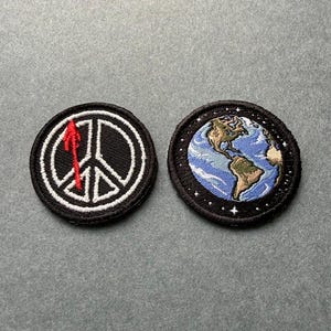 May include: Two circular embroidered patches. The left patch features a white peace symbol on a black background with a vertical red arrow. The right patch displays a detailed globe showing North and South America with white stars on black.