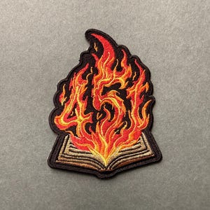 May include: An embroidered patch featuring an open book at the base with flames rising from the pages. The flames are colored in shades of red, orange, and yellow, and they form the number 45. The entire design is outlined in black thread.