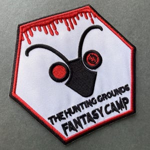 May include: A hexagonal embroidered patch with a white background and a red border featuring a dripping effect. It displays a black insect-like head graphic with red eyes and the black embroidered text THE HUNTING GROUNDS FANTASY CAMP.