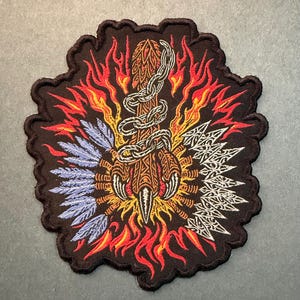 May include: A black embroidered patch featuring a central brown claw wrapped in a silver chain, surrounded by orange and red flames. The left side displays light blue feathers, while the right side features white arrowheads with intricate patterns.
