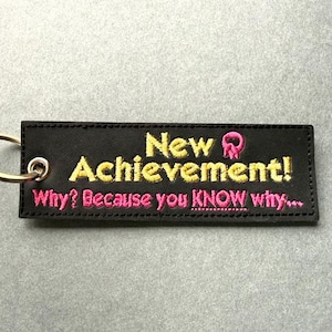 May include: A rectangular black fabric keychain with a silver metal ring. The embroidery features the yellow text New Achievement! with a small pink icon, followed by the pink text Why? Because you KNOW why... stitched on the front surface.
