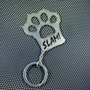 Op de afbeelding: Een zilverkleurige metalen sleutelhanger met een pootafdrukontwerp en het woord "SLAM!" gegraveerd. De sleutelhanger heeft een kleine ketting en een ronde sleutelring.