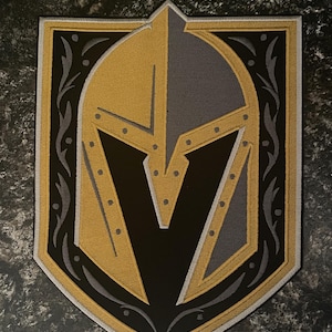 May include: A black, gold, and gray embroidered patch featuring a knight's helmet with a V in the center. The helmet is surrounded by a decorative border.