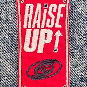 May include: A rectangular red patch with the words "RAISE UP!" embroidered in white. An upward-pointing arrow and a stylized swirl design are also present. The patch is bordered with a tan stitching and is set against a denim background.