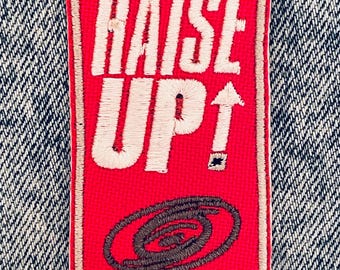以下が含まれることがあります： 「RAISE UP！」の白い刺繍が施された長方形の赤いパッチ。上向きの矢印と渦巻き模様も付いています。パッチはタン色のステッチで縁取られ、デニムの背景に配置されています。