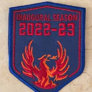 May include: A blue and red embroidered patch with a phoenix design and the text "Inaugural Season 2022-23".
